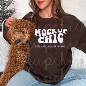 Könnte beinhalten: Ein braunes Sweatshirt mit dem Text "Mock Up Chic" in Weiß, darunter die Worte "Real Models & Real Apparel". Eine Person trägt das Sweatshirt und hält einen braunen Hund.