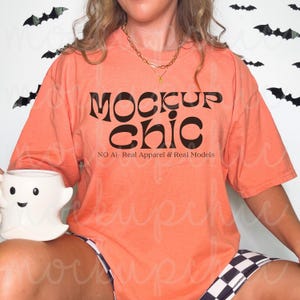 May include: Peach-colored t-shirt with the words "MOCKUP chic" in black, and the text "NO AI- Real Apparel & Real Models". A white ghost-shaped mug is held in the model's lap. Black and white checkered shorts are also visible.