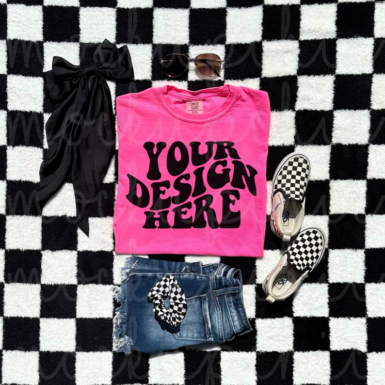 Puede incluir: Camiseta rosa con el texto "YOUR DESIGN HERE" en una fuente retro. La camiseta est&aacute; colocada sobre una superficie a cuadros blancos y negros con un lazo negro, gafas de sol, pantalones cortos de mezclilla azul y zapatos Vans a cuadros blancos y negros.