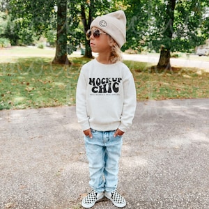 May include: A young person wearing a beige sweatshirt with the text "Mock Up Chic" and the words "Real Models & Real Apparel" printed below. They are also wearing a beige beanie with a smiley face on it and blue jeans.