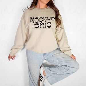 May include: A beige crewneck sweatshirt with the text "MOCKUP chic" in black, with the text "NO AI- Real Apparel & Real Models" below. The sweatshirt is paired with light blue jeans and white sneakers.
