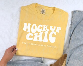 May include: A yellow t-shirt with the text "Mock Up Chic" in white, retro-style font. The text is centered on the shirt and is accompanied by the phrase "Real Models & Real Apparel" below it.