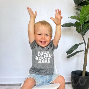 May include: A toddler wearing a heather grey short-sleeved t-shirt with the text "YOUR DESIGN HERE" in white. The toddler is sitting on a white chair with their arms raised in the air.