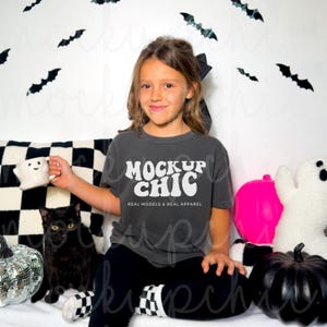 Op de afbeelding: Een kind draagt een antracietkleurig T-shirt met de tekst "Mock Up Chic" in wit. Het shirt is versierd met een retro-lettertype. Het kind zit op een wit oppervlak met een zwart-wit geruit kussen erachter. Er zijn zwarte vleermuizen en een roze pompoen op de achtergrond.