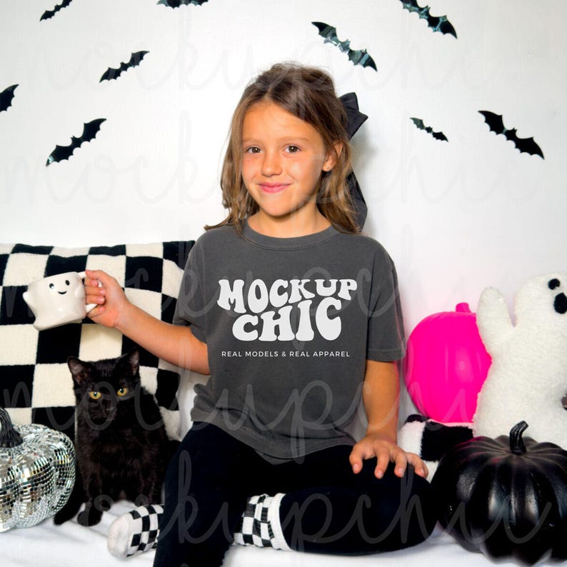 Op de afbeelding: Een kind draagt een antracietkleurig T-shirt met de tekst "Mock Up Chic" in wit. Het shirt is versierd met een retro-lettertype. Het kind zit op een wit oppervlak met een zwart-wit geruit kussen erachter. Er zijn zwarte vleermuizen en een roze pompoen op de achtergrond.