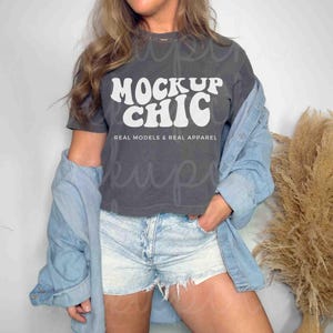 May include: A charcoal gray t-shirt with the text "MOCKUP CHIC" in white, along with the words "REAL MODELS & REAL APPAREL". The model is wearing light wash denim shorts and a light blue denim jacket.