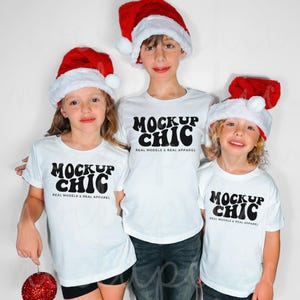 May include: Three children wearing white t-shirts with the text "MOCKUP CHIC REAL MODELS & REAL APPAREL" printed in black. They are wearing red Santa hats.