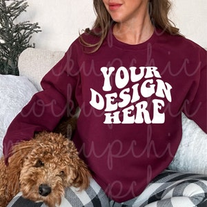 Könnte beinhalten: Ein maroonfarbener Pullover mit dem Text "YOUR DESIGN HERE" in weißer, retro-artiger Schrift. Der Pullover wird von einer Person getragen, die auf einer karierten Decke mit einem braunen Hund sitzt.