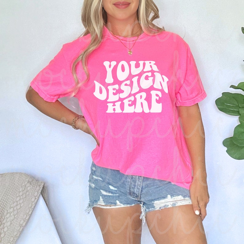 May include: A pink t-shirt with the text "YOUR DESIGN HERE" in white, retro-style lettering. The shirt is oversized and has a relaxed fit.