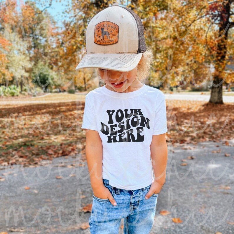 May include: A young child wearing a white t-shirt with the text "YOUR DESIGN HERE" in black. The child is also wearing a brown and black baseball cap with a patch that says "LOCALBOY OUTFITTERS" and blue jeans.