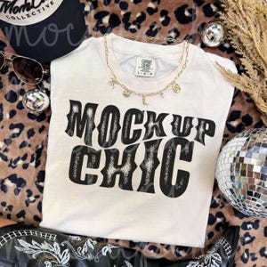 May include: Cream-colored t-shirt with the words "MOCKUP CHIC" in a black and white design. A gold necklace with charms and sunglasses are on the shirt. A black hat and a disco ball are also in the image.
