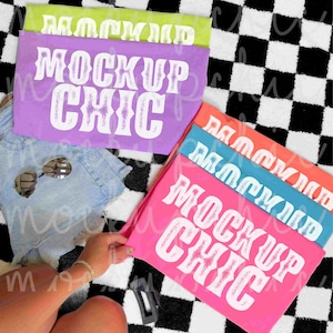 May include: A stack of colorful t-shirts with the text "MOCKUP CHIC" in a bold, white font. The shirts are in shades of pink, purple, green, and blue. Sunglasses and denim shorts are also visible. The background is a black and white checkered pattern.