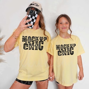 May include: Two people wearing matching light yellow t-shirts with the text "MOCKUP CHIC" in black. One person is wearing a baseball cap and holding a phone with a black and white checkered case. The other person is a child.