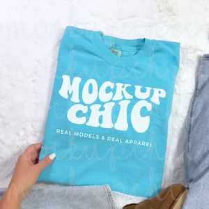 May include: A light blue t-shirt with the text "Mock Up Chic" in white, retro-style lettering. The text is centered on the shirt and is followed by the phrase "Real Models & Real Apparel".