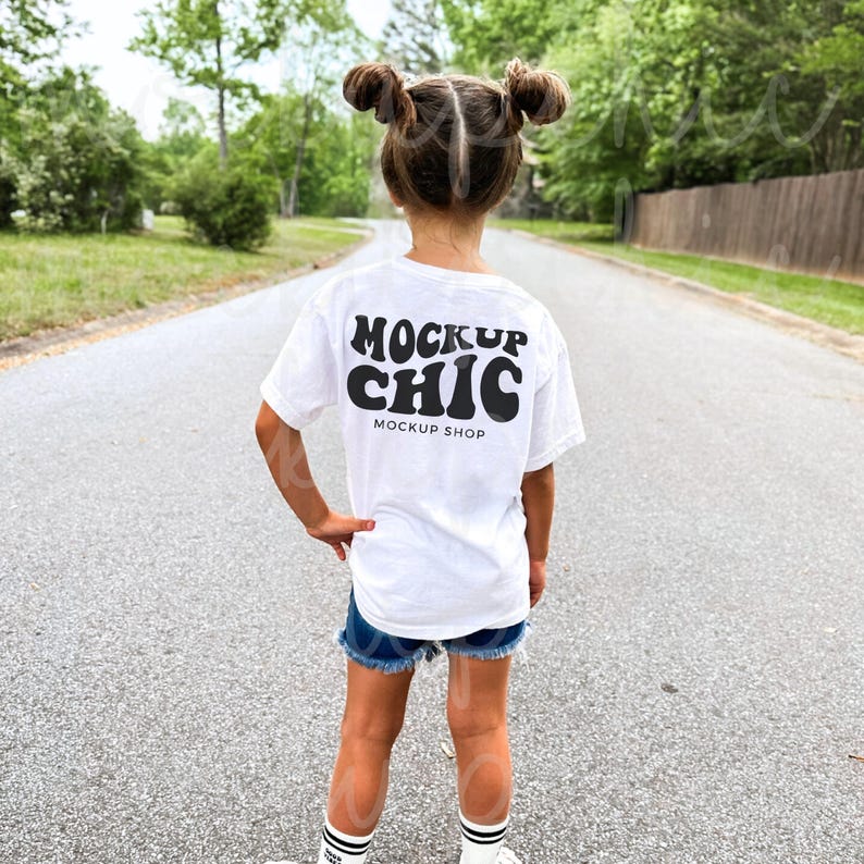 May include: A white t-shirt with black text that reads "Mockup Chic" and "Mockup Shop" on the back. The shirt is being worn by a child.