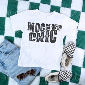 Könnte beinhalten: Weißes T-Shirt mit schwarzem Text, der "Mock Up Chic" auf einem grün-weiß karierten Hintergrund sagt. Das T-Shirt liegt auf einer blauen Jeans und karierten Vans-Schuhen.