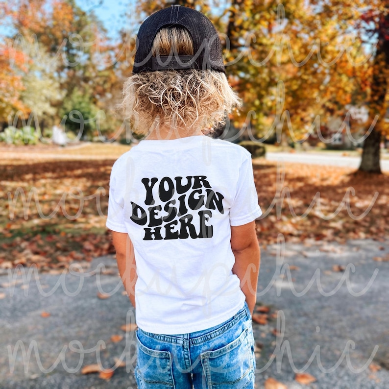 May include: A white t-shirt with the text "YOUR DESIGN HERE" in black, worn by a child with curly blonde hair and a black baseball cap. The child is standing in front of a background of fall foliage.