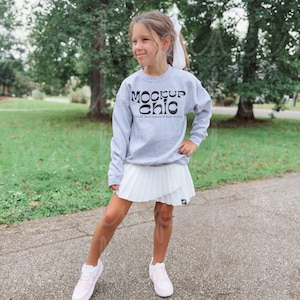 May include: A young person wearing a gray sweatshirt with the text "MOCKUP chic" and a white pleated skirt. The outfit is completed with white and pink sneakers. The background features green trees and a paved path.