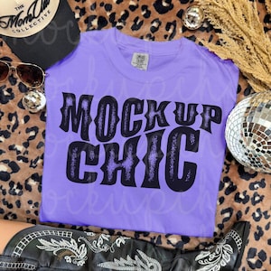 May include: A lilac-colored t-shirt with the text "MOCK UP CHIC" in a black, retro font. The shirt is displayed with a black hat, sunglasses, and a pair of black cowboy boots. Decorative disco balls and dried wheat are also present.