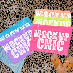 May include: A collection of colorful t-shirts with the text "MOCKUP CHIC" in white. The shirts are in shades of pink, orange, green, blue, and purple. The background features a leopard print pattern and a pair of brown sandals.