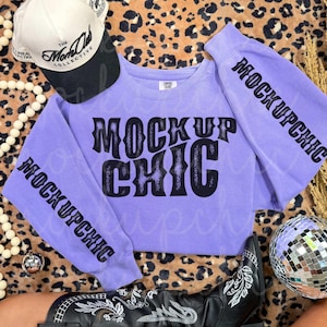May include: A lavender sweatshirt with the text "MOCK UP CHIC" in black lettering. The sleeves also have the text "MOCK UP CHIC" printed on them. A white and black baseball cap and black cowboy boots are also in the image.