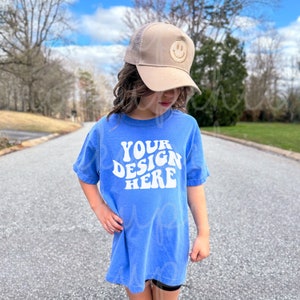 May include: A child wearing a blue t-shirt with the text "YOUR DESIGN HERE" in white. The child is also wearing a brown mesh baseball cap with a smiley face.
