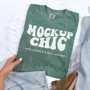 May include: A green t-shirt with the text "MOCKUP CHIC" in white, retro-style lettering. The text is centered on the shirt and is surrounded by the words "REAL MODELS & REAL APPAREL".