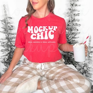 May include: A red t-shirt with the text "Mock Up Chic" in white, with the words "Real Models & Real Apparel" underneath. The person is wearing plaid pajama pants and holding a mug with marshmallows.