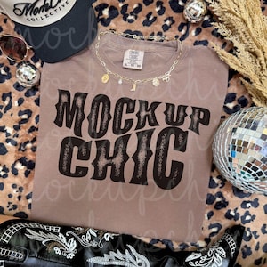 May include: A brown t-shirt with the text "MOCKUP CHIC" in a bold, distressed font. The shirt is paired with a gold necklace, a black and white baseball cap, sunglasses, and a pair of black cowboy boots. A disco ball and leopard print fabric are also visible.