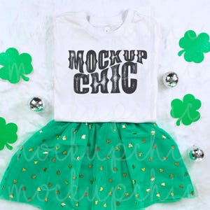 Puede incluir: Camiseta blanca con el texto "Mock Up Chic" en letras negras. La camiseta se combina con una falda de tul verde con tréboles de purpurina dorada.