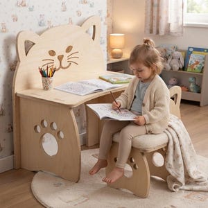 May include: A light-colored wooden desk and matching stool set featuring a cat-shaped backrest and paw-print cutouts on the side panels. The desk surface holds a cup of colored pencils and an open coloring book, while the stool has a cushioned seat.