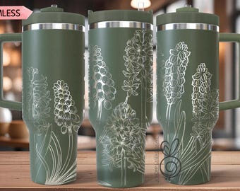 May include: Three olive green insulated tumblers with silver floral designs. Each tumbler has a handle and a lid. The floral design is a detailed outline of hyacinth flowers. The tumblers are on a wooden surface.