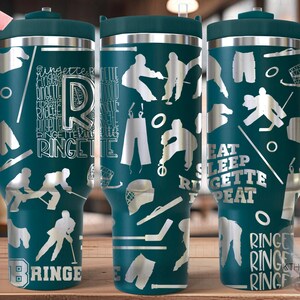 May include: A teal stainless steel travel mug with a white handle. The mug has a white design of hockey players and the words "Eat Sleep Ringette Repeat" on it.