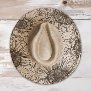 May include: A beige fedora hat with a sunflower design. The hat features a wide brim with a detailed floral pattern in shades of brown and tan. The crown of the hat is a solid beige color. The hat is displayed on a light-colored wooden surface.