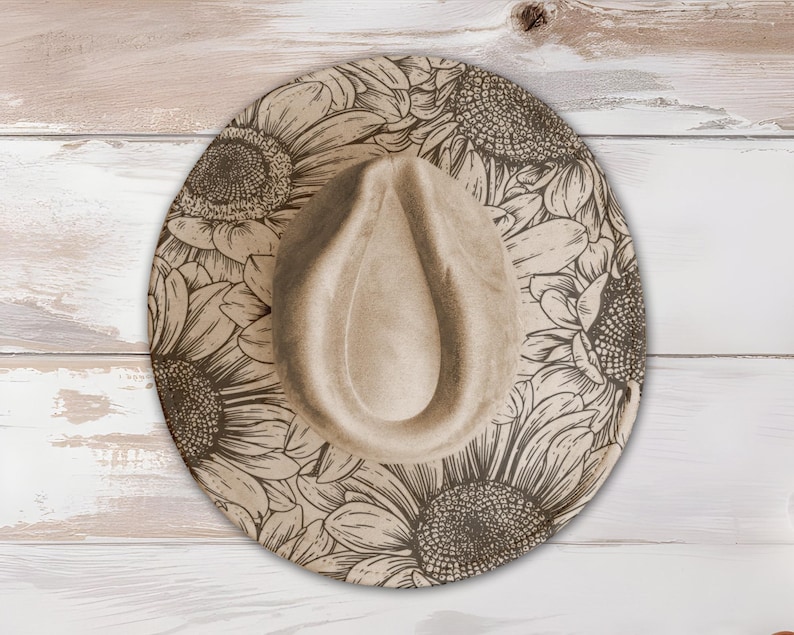 May include: A beige fedora hat with a sunflower design. The hat features a wide brim with a detailed floral pattern in shades of brown and tan. The crown of the hat is a solid beige color. The hat is displayed on a light-colored wooden surface.