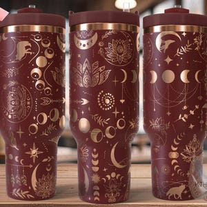 Pode incluir: Três copos de aço inoxidável com base bordô e detalhes dourados. Os copos apresentam um padrão repetitivo de luas, estrelas e desenhos florais. Os copos têm tampas e alças. O texto "SEAMLESS" é visível no canto superior esquerdo da imagem.