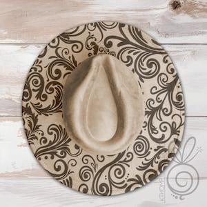 May include: A beige fedora hat with a brown, swirling, floral pattern. The hat has a wide brim and a teardrop-shaped crown. The hat is set against a light wood background.