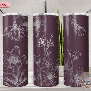 May include: Three stainless steel tumblers with a purple background and white floral designs. The tumblers are arranged in a row, with the middle tumbler slightly in front of the other two. The tumblers are all the same size and shape, and they have a lid and a straw.