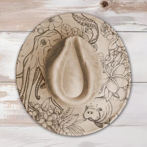 May include: A beige fedora hat with a wide brim, featuring a detailed black line drawing of an octopus, flowers, and a manatee. The hat has a teardrop-shaped crown and a structured design, suitable for sun protection and fashion.
