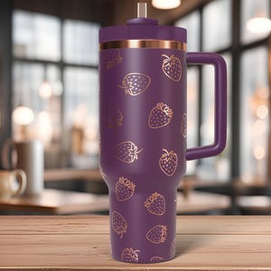 May include: A large, purple insulated tumbler with a handle and a straw. The tumbler is decorated with gold-colored strawberry illustrations. The top and handle are a darker shade of purple, and the rim is a copper color.
