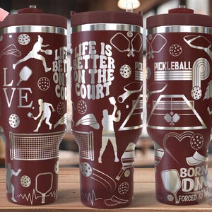 May include: Three stainless steel tumblers with a maroon exterior and silver engraved designs. The tumblers feature pickleball-themed graphics, including paddles, nets, and the text "Life is better on the court", "Pickleball", and "Born to be forced to play".
