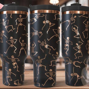 Könnte beinhalten: Drei schwarze Edelstahlbecher mit kupferfarbenem Deckel und Henkel. Die Becher sind mit einem sich wiederholenden Muster aus goldenen Skeletten verziert, die auf schwarzem Hintergrund tanzen. Die Becher sind nahtlos.