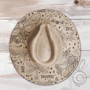 May include: Tan fedora hat with intricate black line art. The hat features illustrations of butterflies, flowers, and a dragonfly, along with the words "MAGIC", "LUCK", "LOVE", and "PASSION". The hat has a classic teardrop crown and a wide brim.