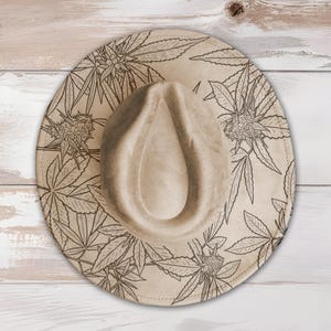 May include: A tan fedora-style hat with a wide brim. The hat features a repeating pattern of cannabis leaves and flowers in a dark brown color. The hat is made of a soft material and is suitable for sun protection.
