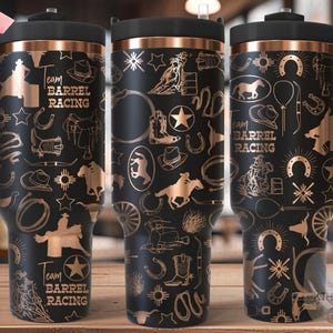 May include: Black tumbler with a copper-colored design featuring cowboy and barrel racing motifs. The tumbler has a black handle and lid, and the text "Team Barrel Racing" is visible.