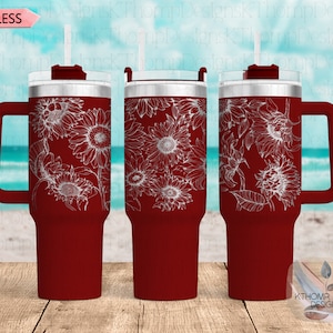 Puede incluir: Un vaso de acero inoxidable rojo con un diseño de girasol blanco. El vaso tiene un asa y una tapa con un agujero para pajita. El vaso se muestra sobre una superficie de madera con un fondo de playa.