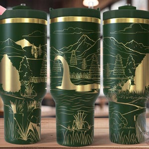 May include: A green and gold stainless steel travel mug featuring a design of a lake monster in a woodland setting. The mug has a handle and a lid.