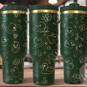 Könnte beinhalten: Ein grüner Edelstahlbecher mit goldenem Deckel und Henkel. Der Becher hat ein nahtloses Design mit einem sich wiederholenden Muster aus goldenen Gummienenten auf grünem Hintergrund.