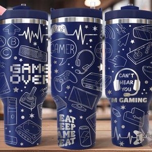 May include: A blue stainless steel travel mug with a white gamer-themed design. The design includes a video game controller, a headset, and the text "Game Over", "Gamer", "Can't Hear You", and "I'm Gaming".