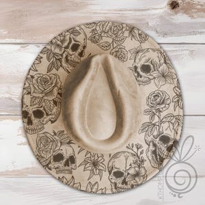 Op de afbeelding: Een beige fedora-hoed met een brede rand. De rand is versierd met een herhalend patroon van schedels en rozen in donkerbruin. De hoed heeft een klassieke druppelvormige kroon.
